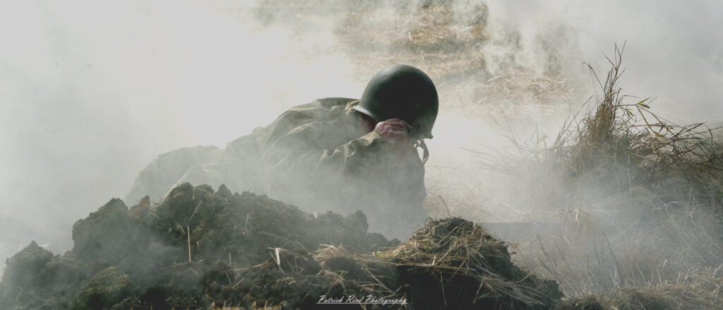 "Image of a soldier standing resolutely in a smoky field, surrounded by a haze of battle. The soldier, dressed in a WWII-era uniform, grips his rifle tightly, scanning the environment with a determined expression. The smoke curls around him, creating a dramatic atmosphere that highlights the chaos of war. The background features silhouettes of trees and hints of distant action, emphasizing the soldier's isolation and bravery in a challenging situation."